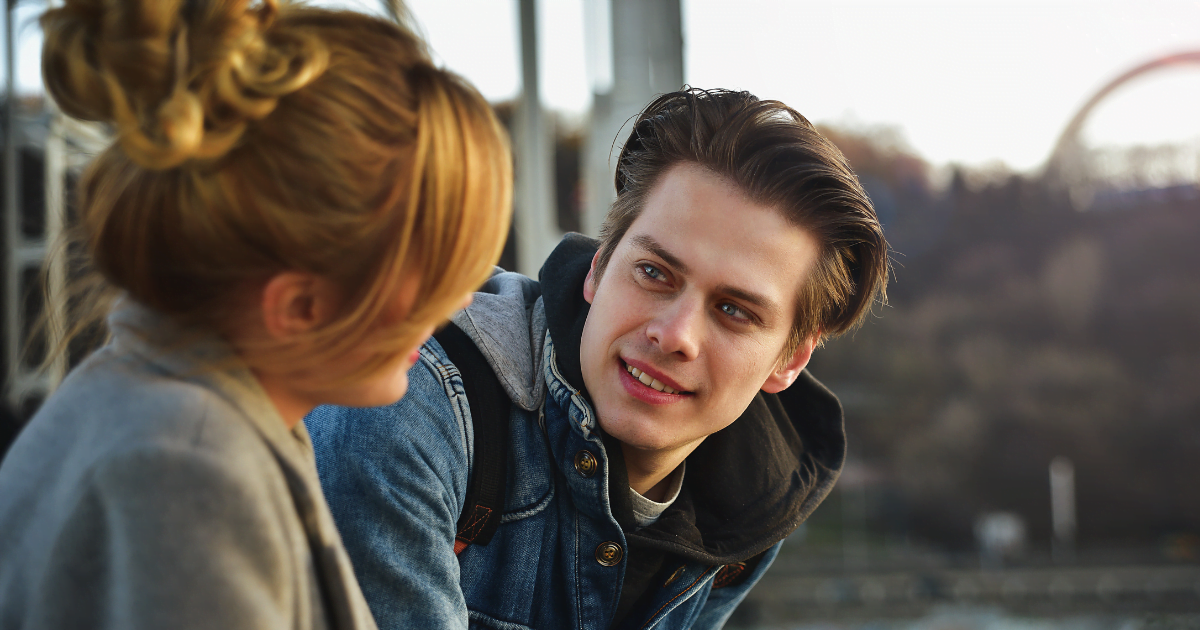 A young man gazes affectionately at a woman, radiating happiness and affection, showcasing the beauty of love languages in an outdoor setting with a bridge and trees in the background during sunset.