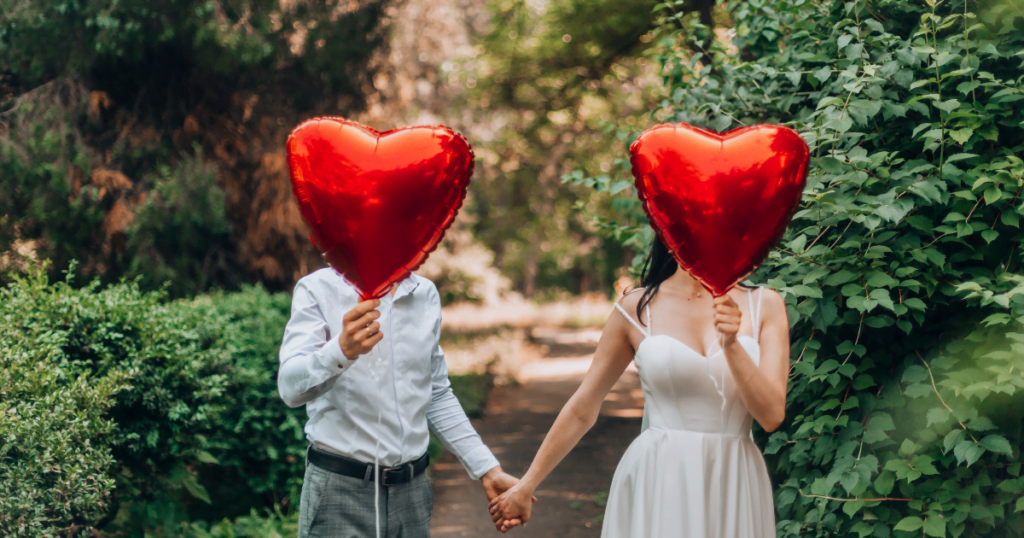 A couple holding hands, each covering their face with a red heart-shaped balloon, walking down a pathway surrounded by lush greenery, showing love and care in their nurturing relationship.