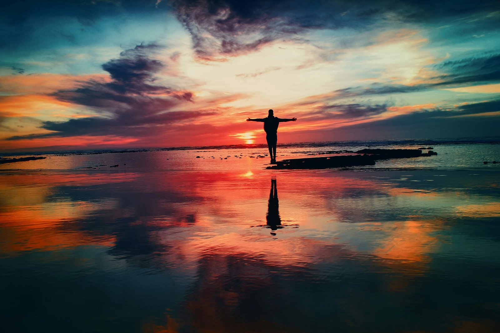 A person standing on a beach at sunset, with their arms raised towards the sky, represents connecting with the Laws of Energy in the universe.