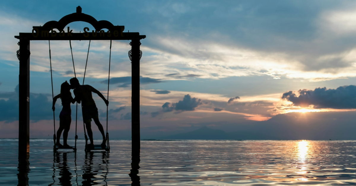 A romantic couple kissing on a swing in the water, surrounded by love.